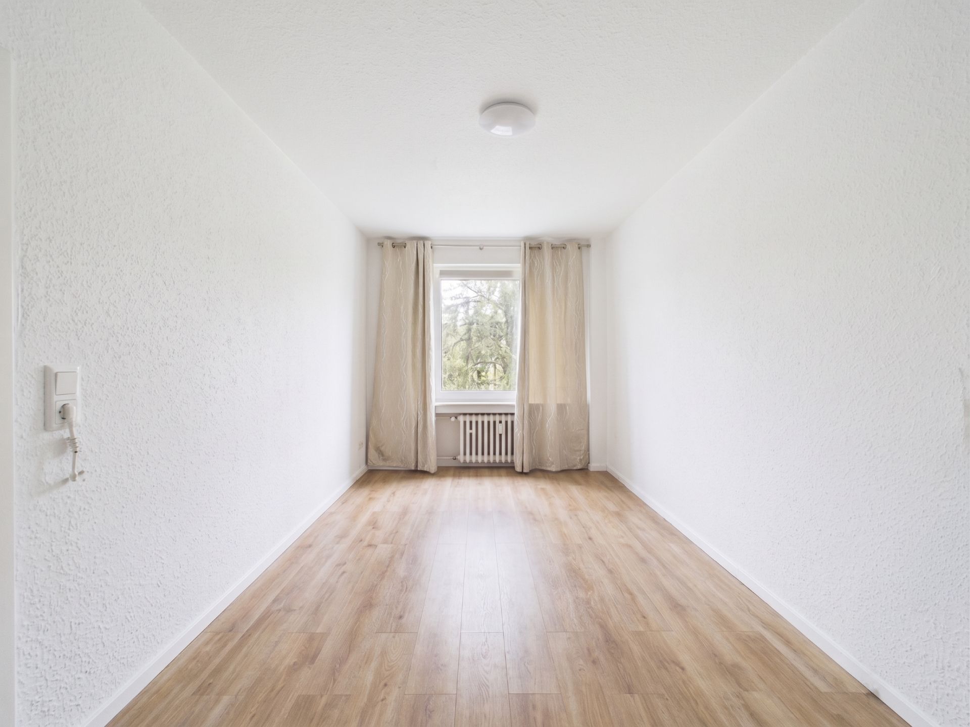 Leerer, heller Raum mit Holzfußboden, weißem Wänden, Fenster, Vorhängen und Heizkörper.