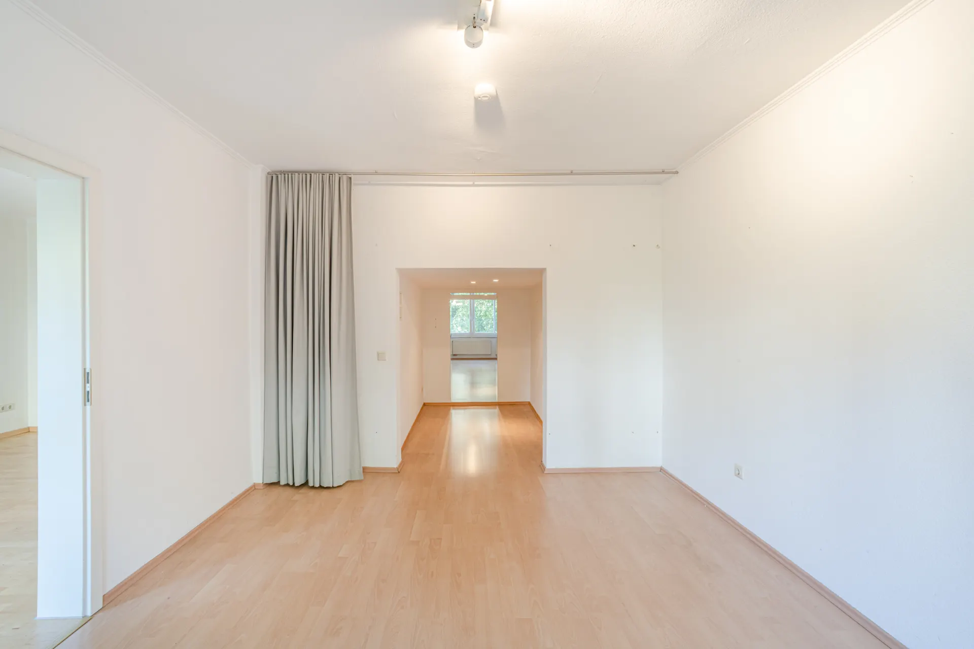 Heller leerer Raum mit Holzfußboden, weißen Wänden und großem Fenster im Hintergrund.