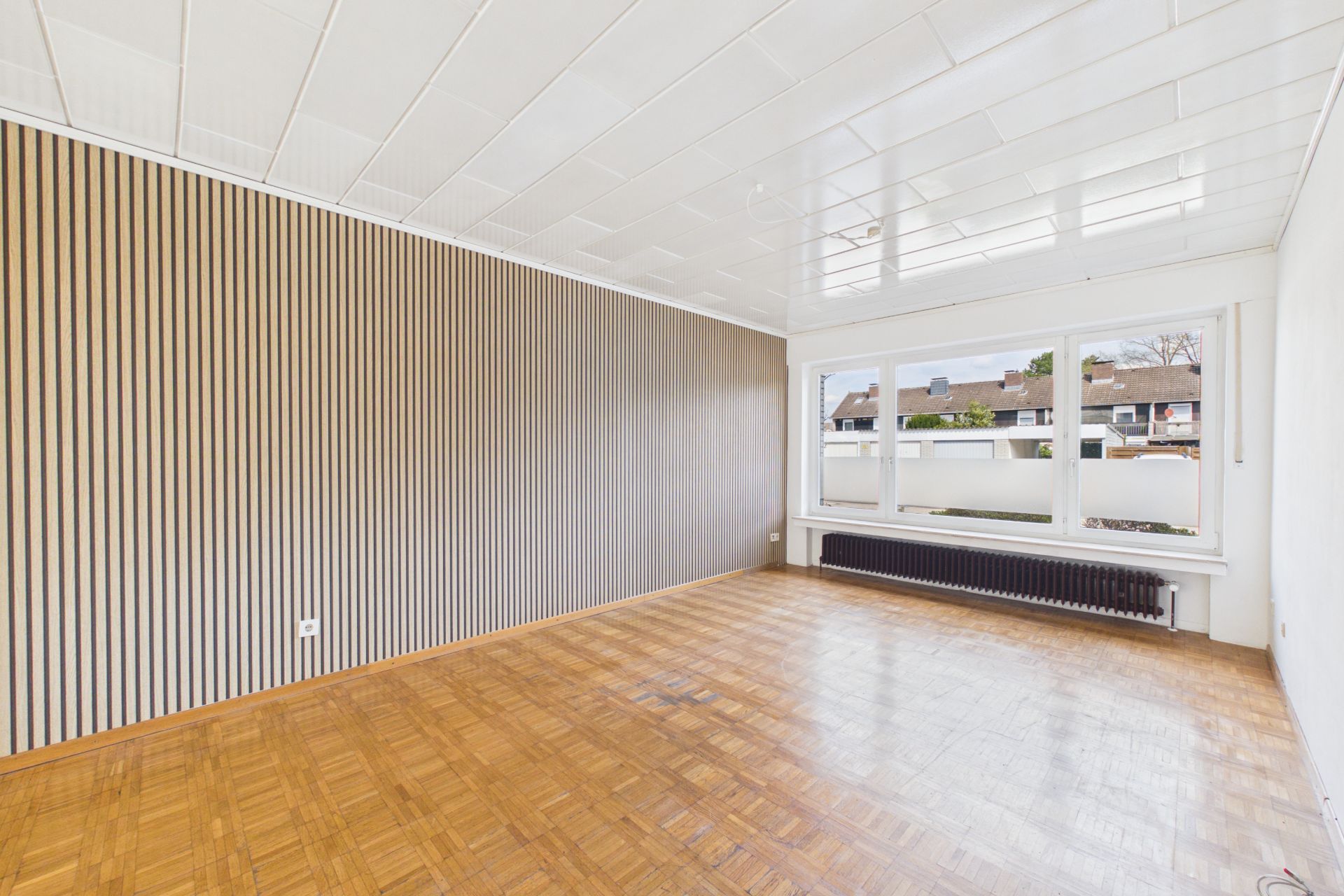 Heller, leerer Raum mit Holzfußboden, großer Fensterfront und gestreifter Wandverkleidung.