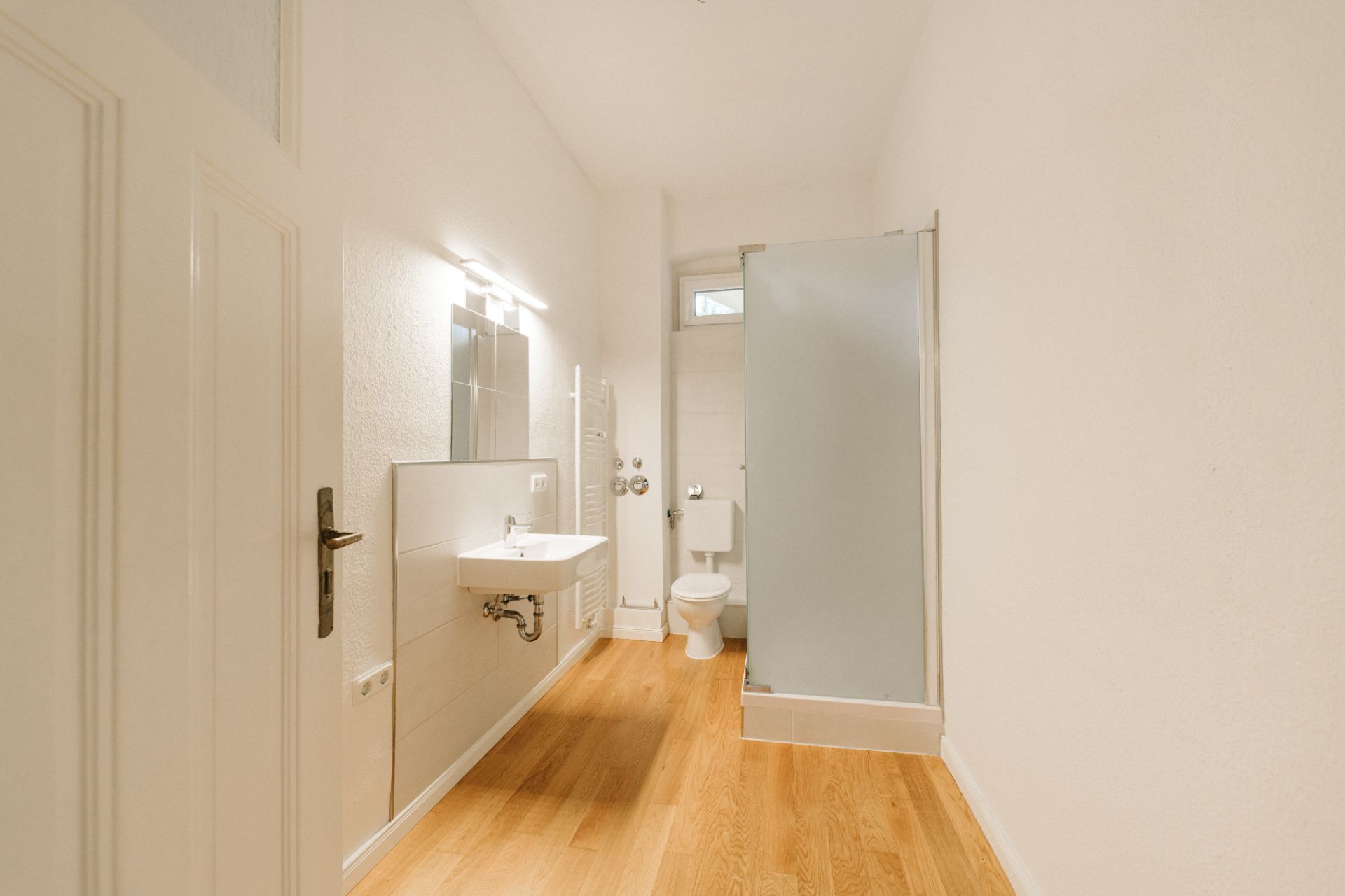 Helles Badezimmer mit Holzboden, Waschbecken, Spiegel, Toilette und Dusche mit Milchglaswand.