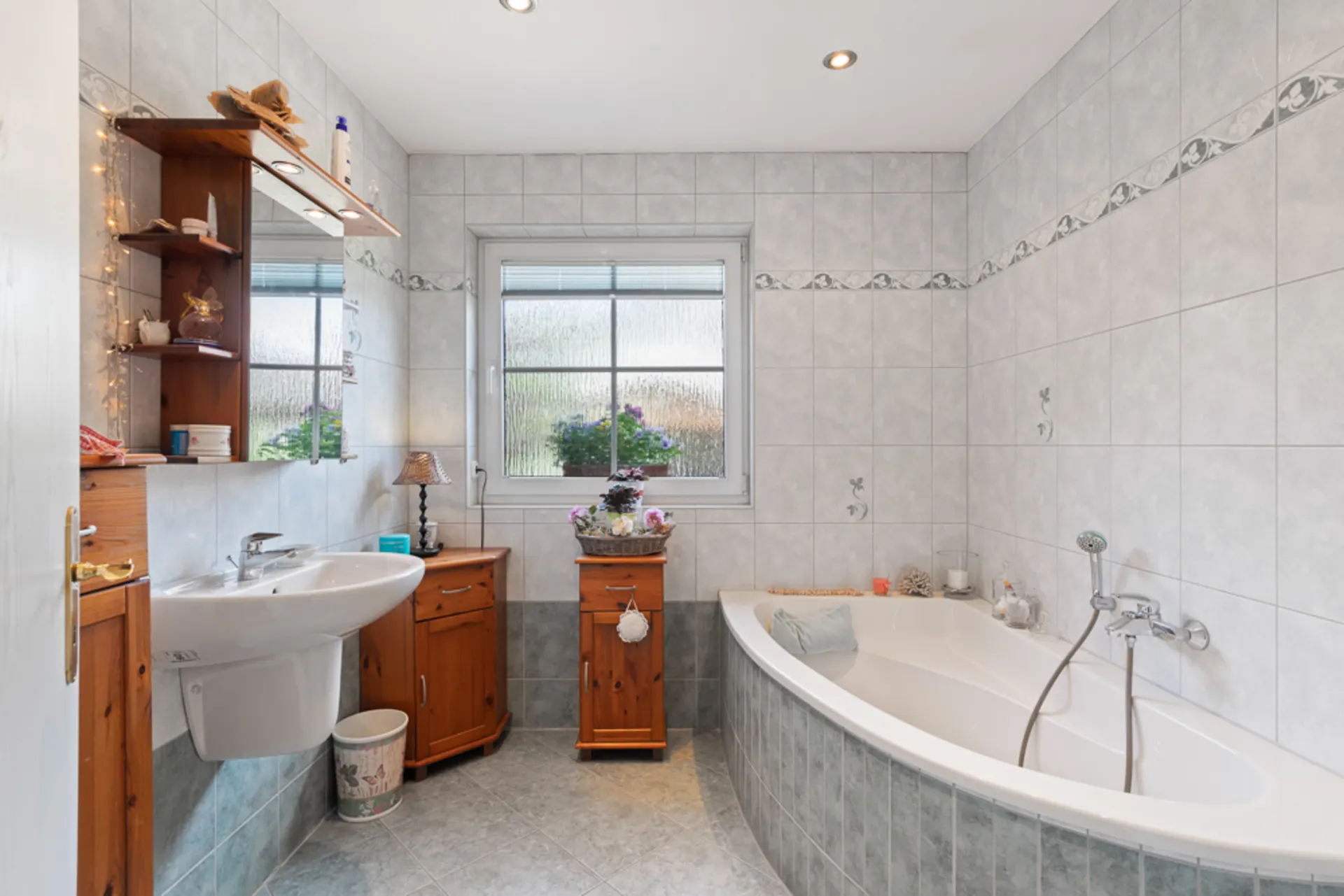 Helles Badezimmer mit Eckbadewanne, Waschbecken, Holzschränken und großem Fenster.