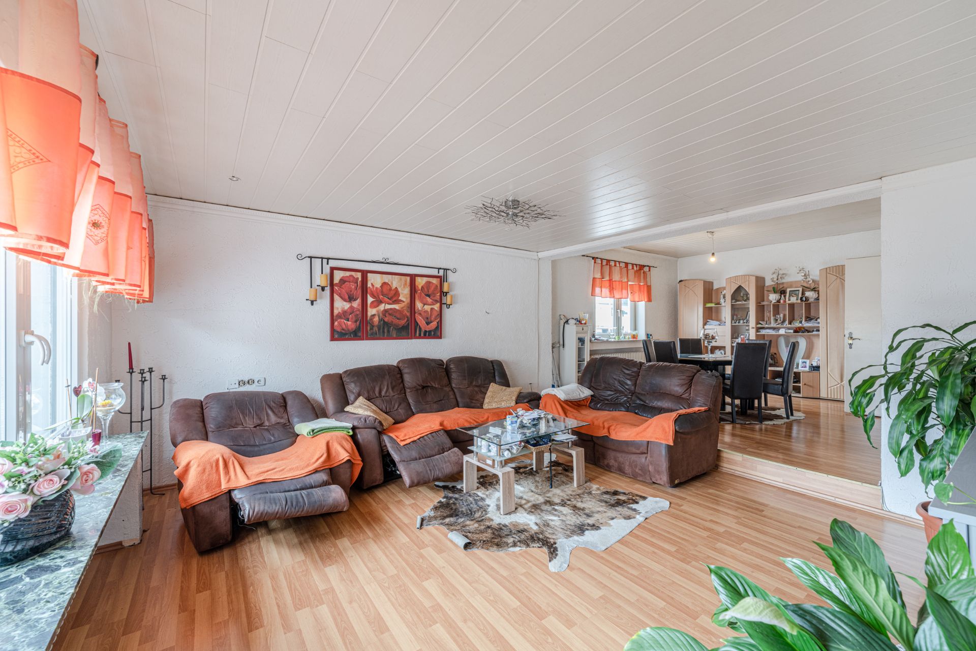 Wohnzimmer mit braunen Sofas, Couchtisch, orangefarbenen Vorhängen und offener Essbereich im Hintergrund.