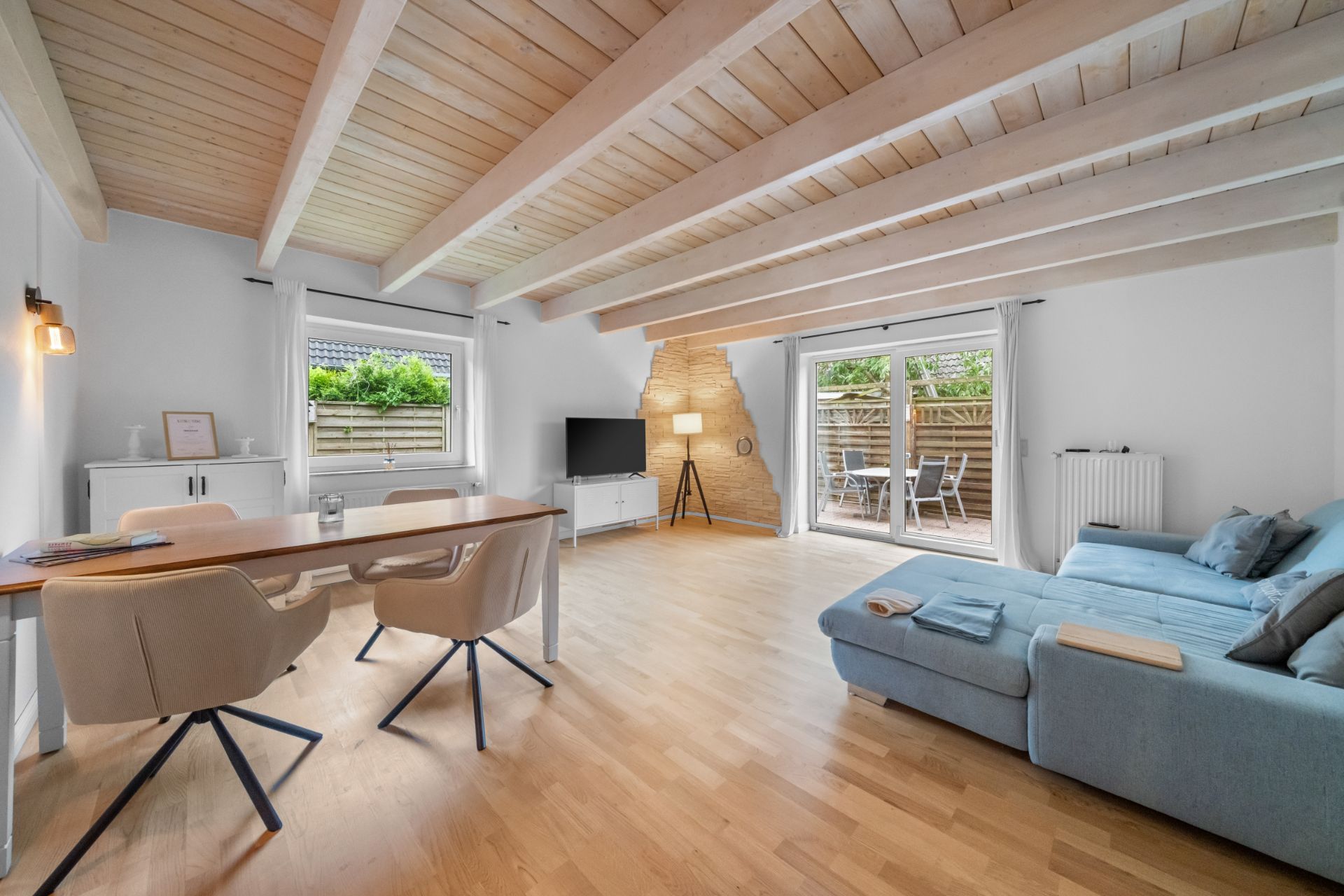 Helles Wohnzimmer mit Holzbalkendecke, Sofa, Esstisch, Fernseher und Zugang zur Terrasse.