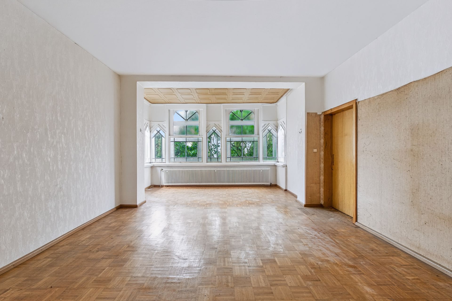 Leeres Zimmer mit Parkettboden, großen Fenstern und Holztür auf der rechten Seite.