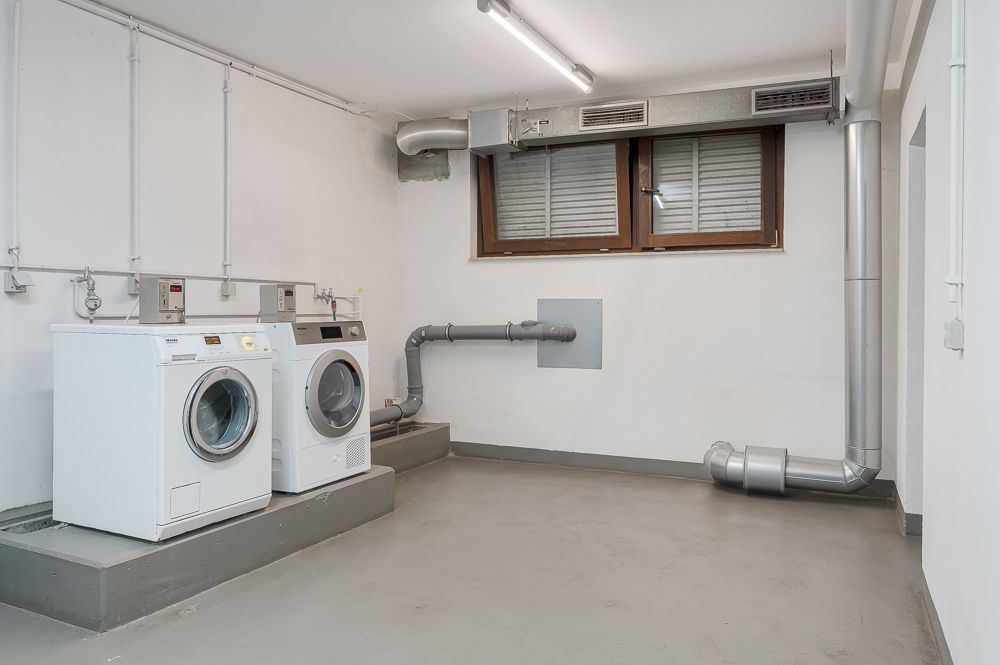 Kellerraum mit zwei Waschmaschinen, Lüftungsrohren und einem Fenster mit Rollladen.