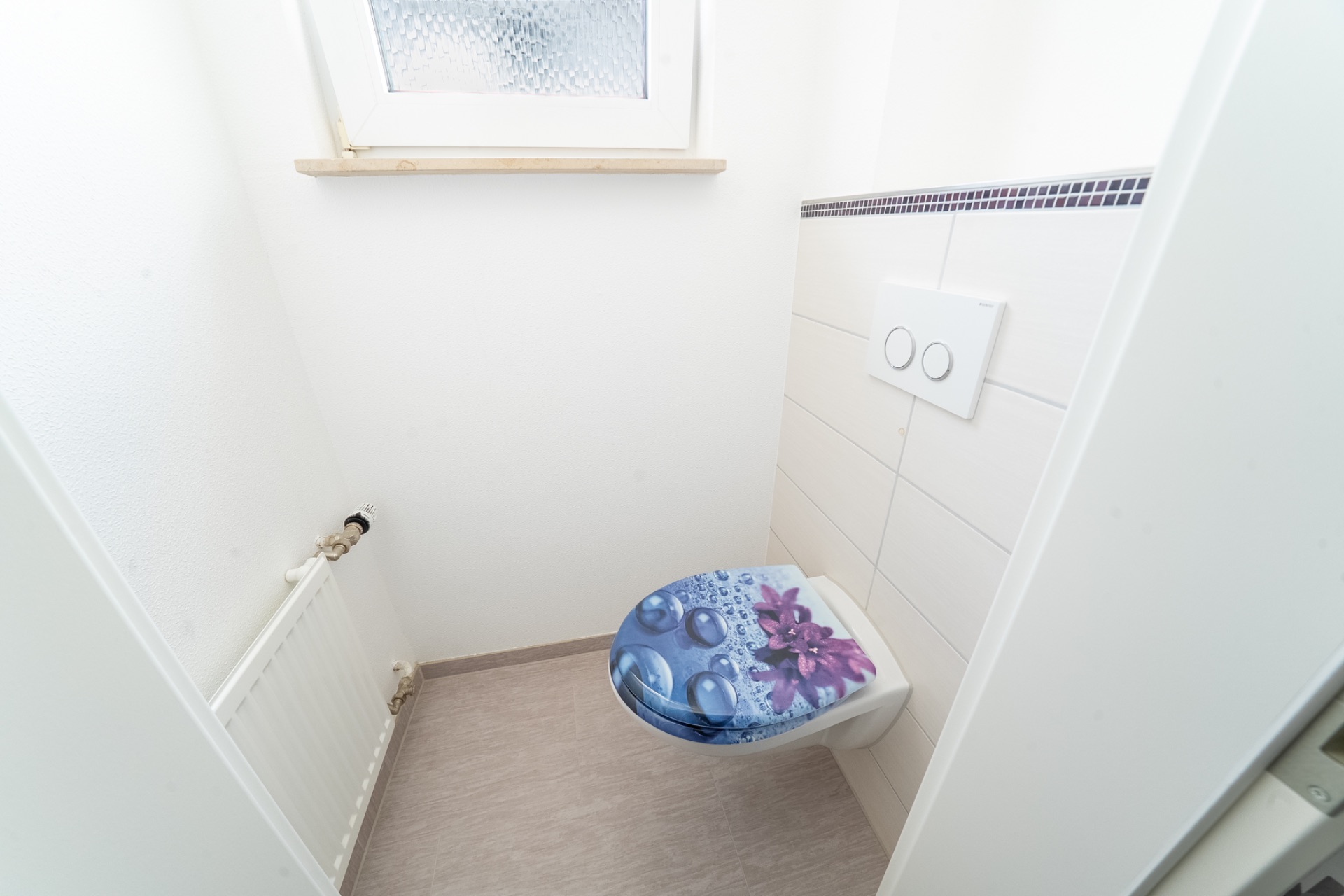 Kleines WC mit Fenster, Heizkörper und Toilettendeckel mit Blütenmuster.
