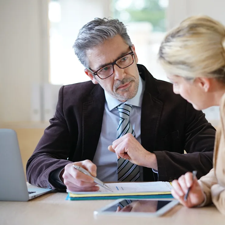 Zwei Personen sitzen am Tisch und sprechen, mit Papieren und Laptop vor sich.