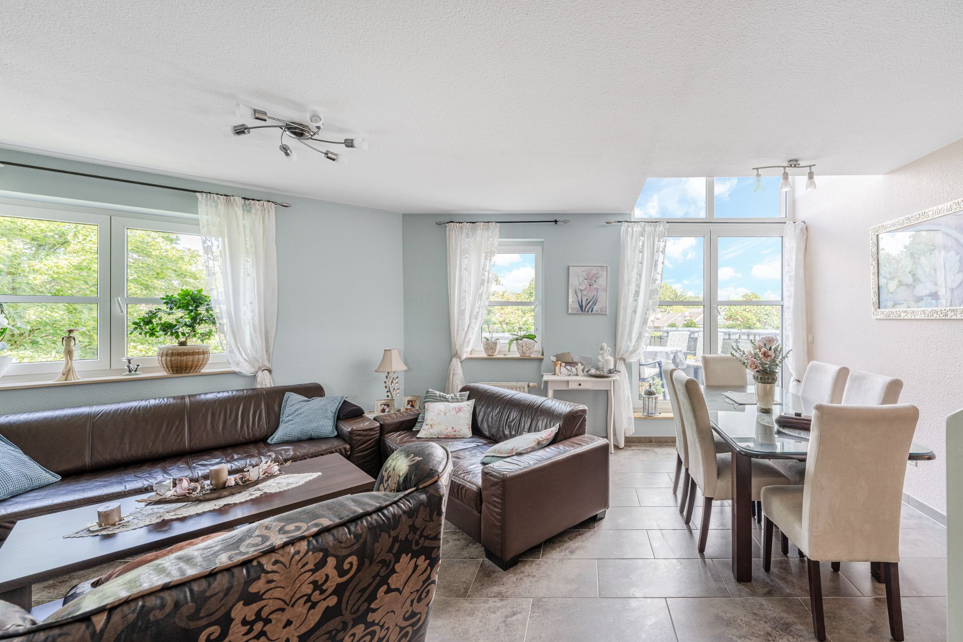 Helles Wohnzimmer mit braunen Sofas, Esstisch mit Stühlen und großen Fenstern mit weißen Vorhängen.