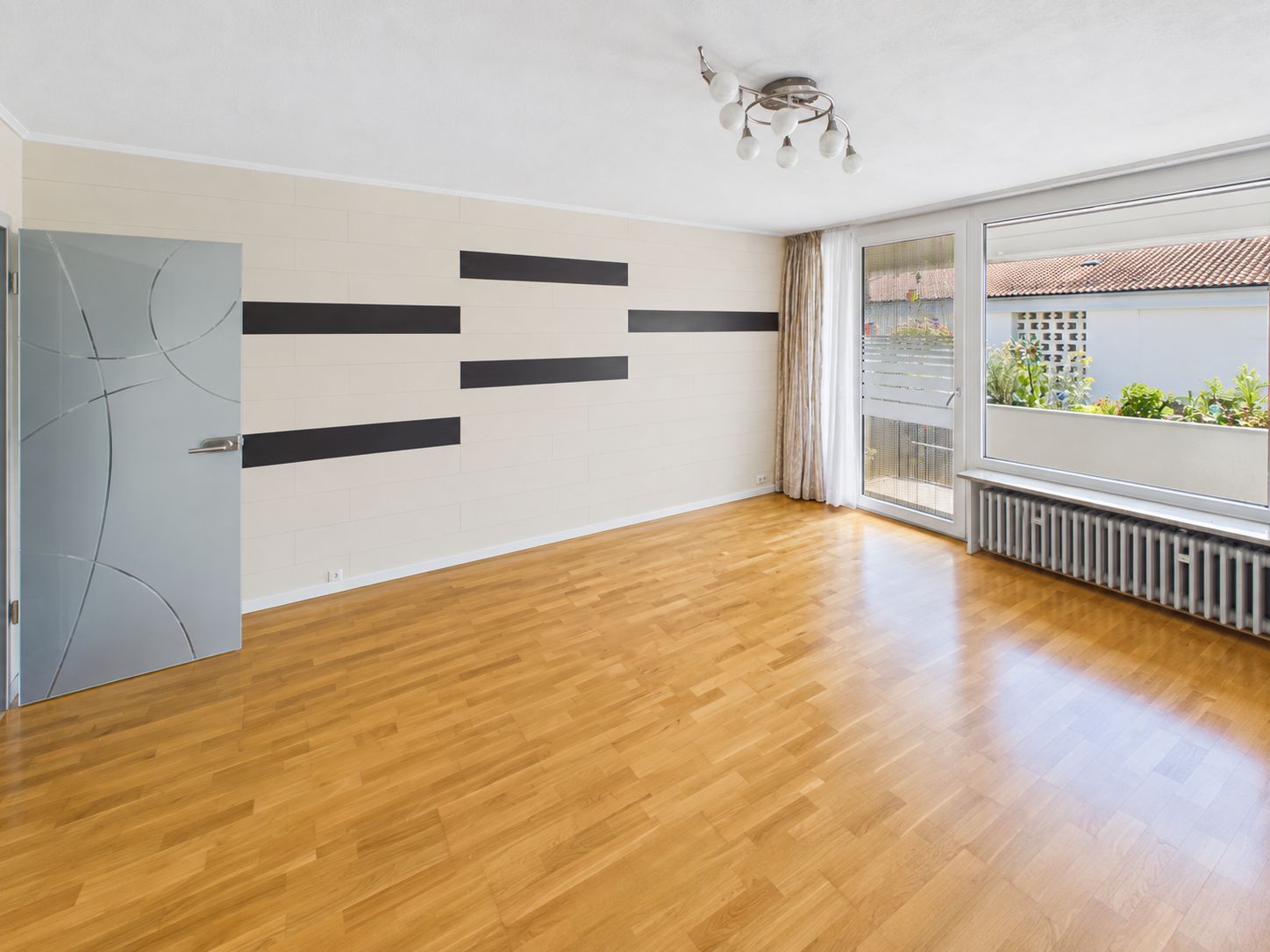 Helles, leeres Zimmer mit Holzboden, Glas­tür, Heizkörper und großen Fenstern zur Terrasse.