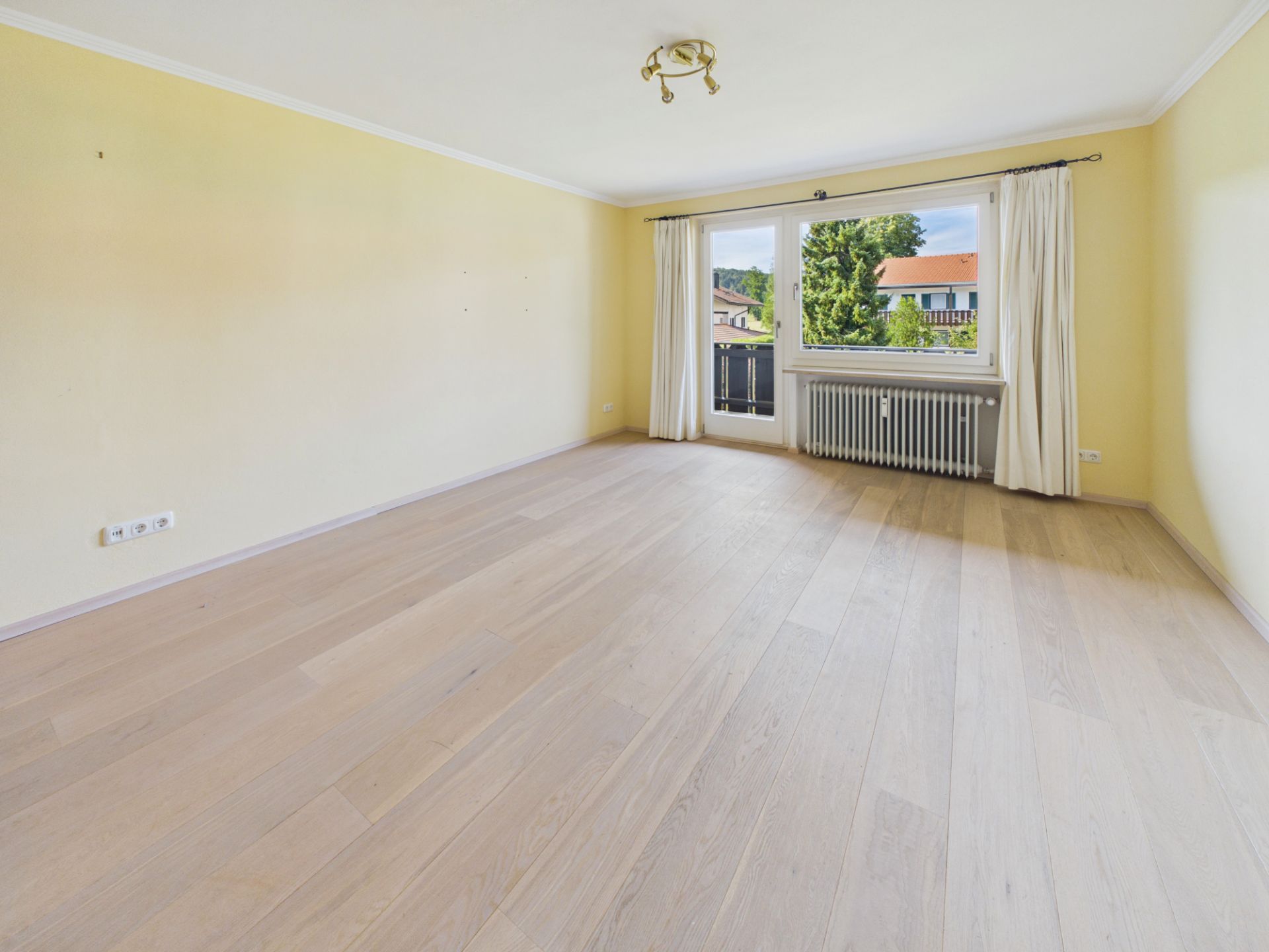 Helles, leeres Zimmer mit Holzfußboden, gelben Wänden und großem Fenster mit Vorhängen.
