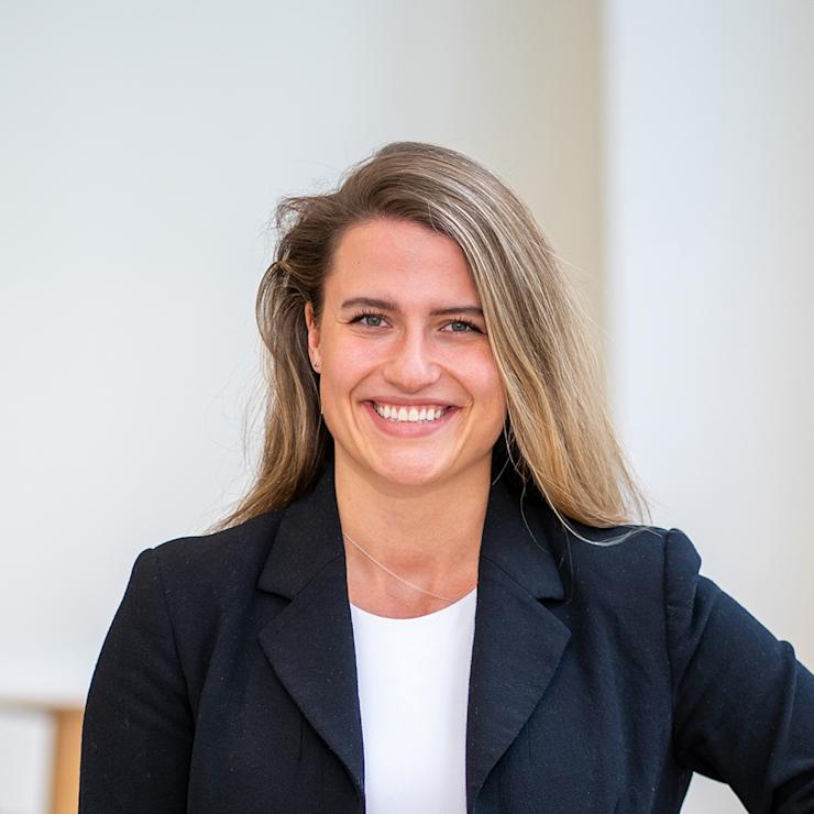 Eine Frau mit langen Haaren und schwarzem Blazer steht lächelnd vor hellem Hintergrund.