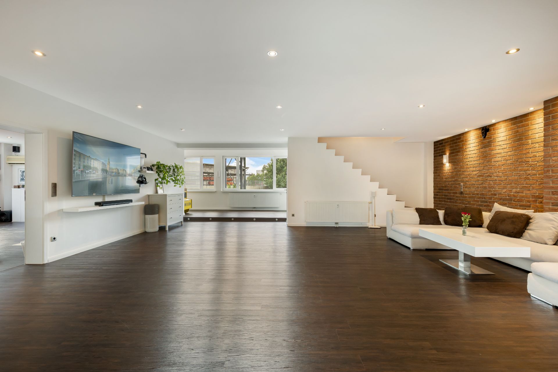 Großes Wohnzimmer mit dunklem Holzboden, Ecksofa, Fernseher, Ziegelwand und Treppe.