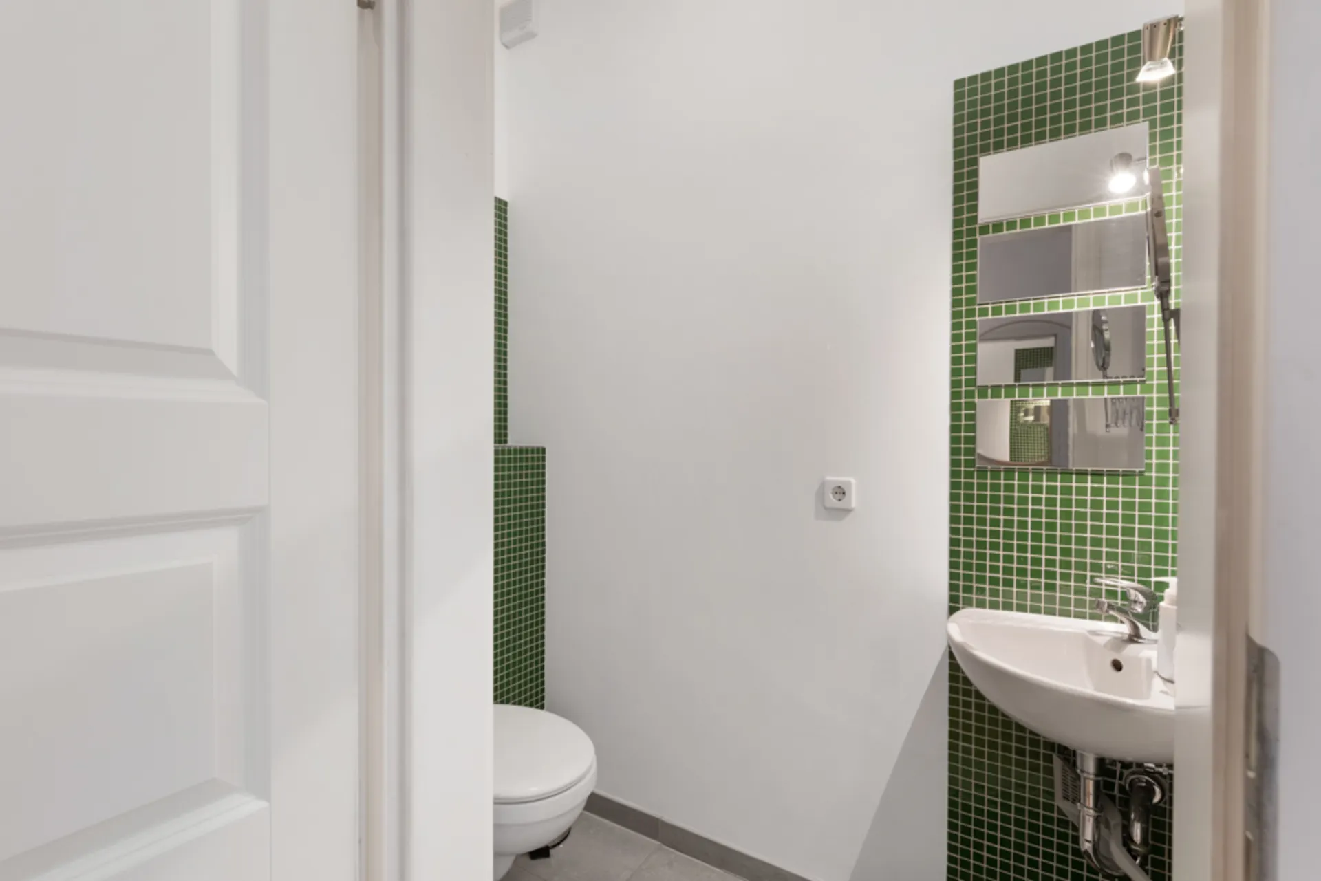Kleines Badezimmer mit grünen Fliesen, Wandspiegeln, Waschbecken und Toilette.