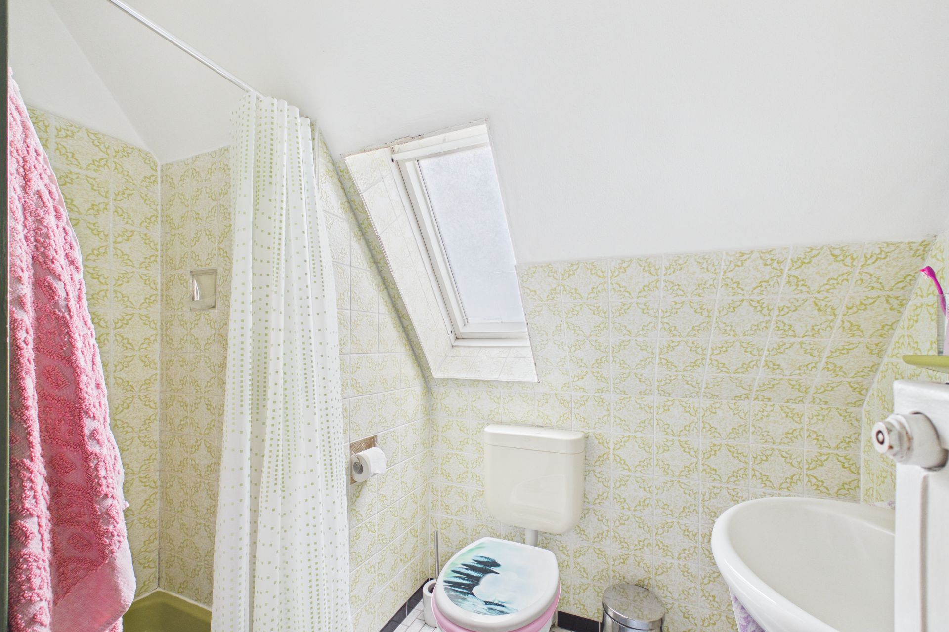 Kleines Bad mit Toilette, Waschbecken, Dusche, Fenster und gemusterten Fliesen.