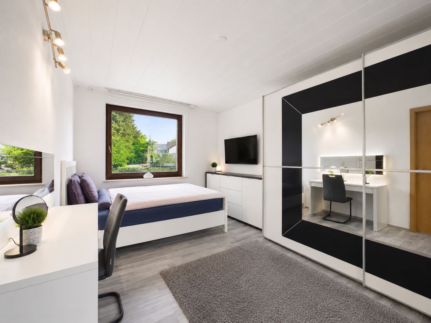 Modernes Schlafzimmer mit Bett, Schreibtisch, großem Spiegel und Fenster mit Blick ins Grüne.