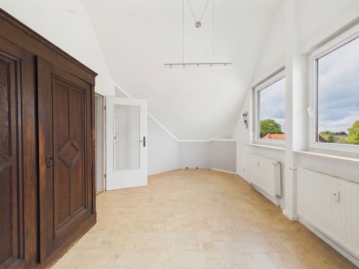 Helles Zimmer mit schräger Decke, zwei Fenstern, Heizkörper und großem Holzschrank.