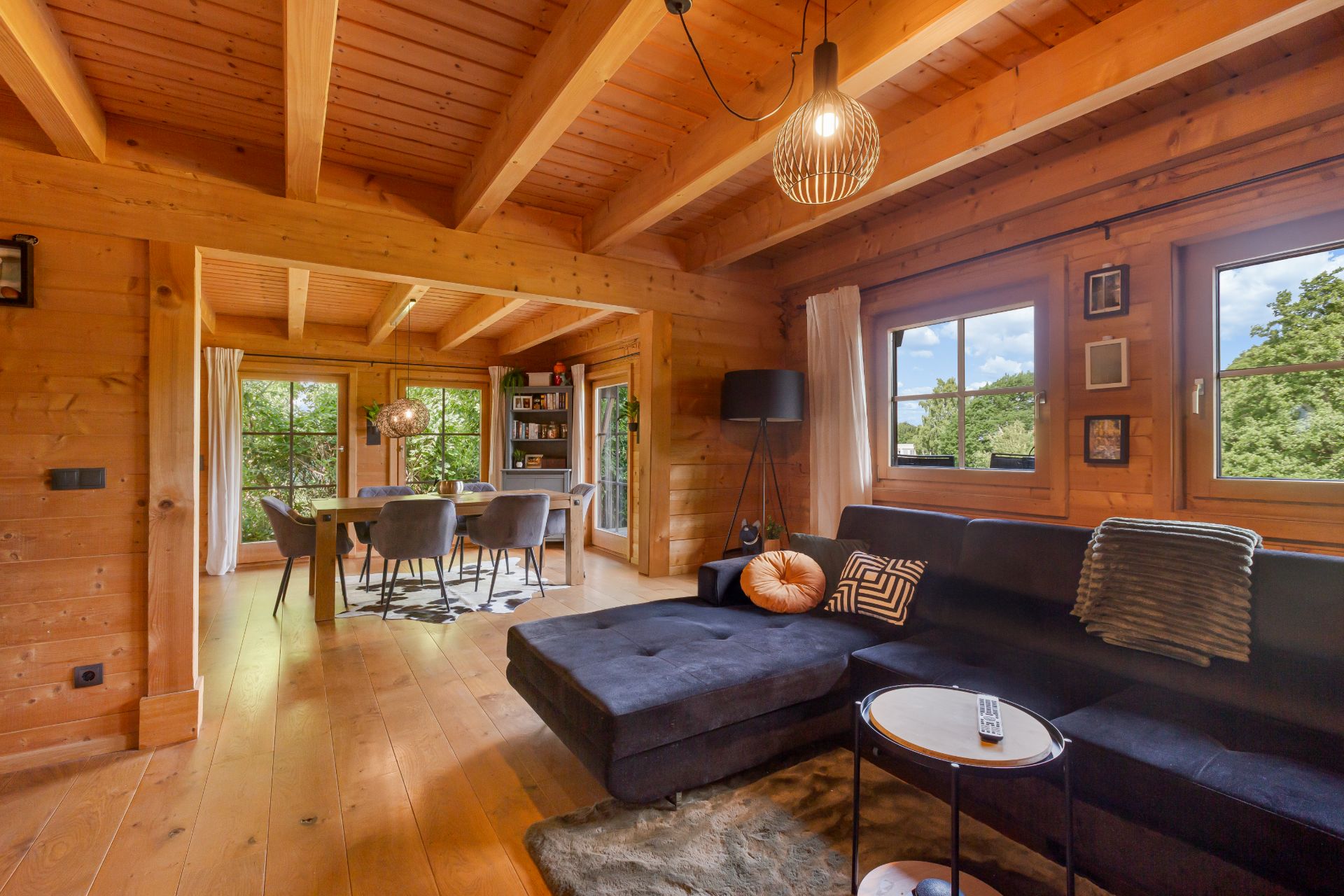 Wohnzimmer aus Holz mit Ecksofa, Esstisch und großen Fenstern, Blick ins Grüne.