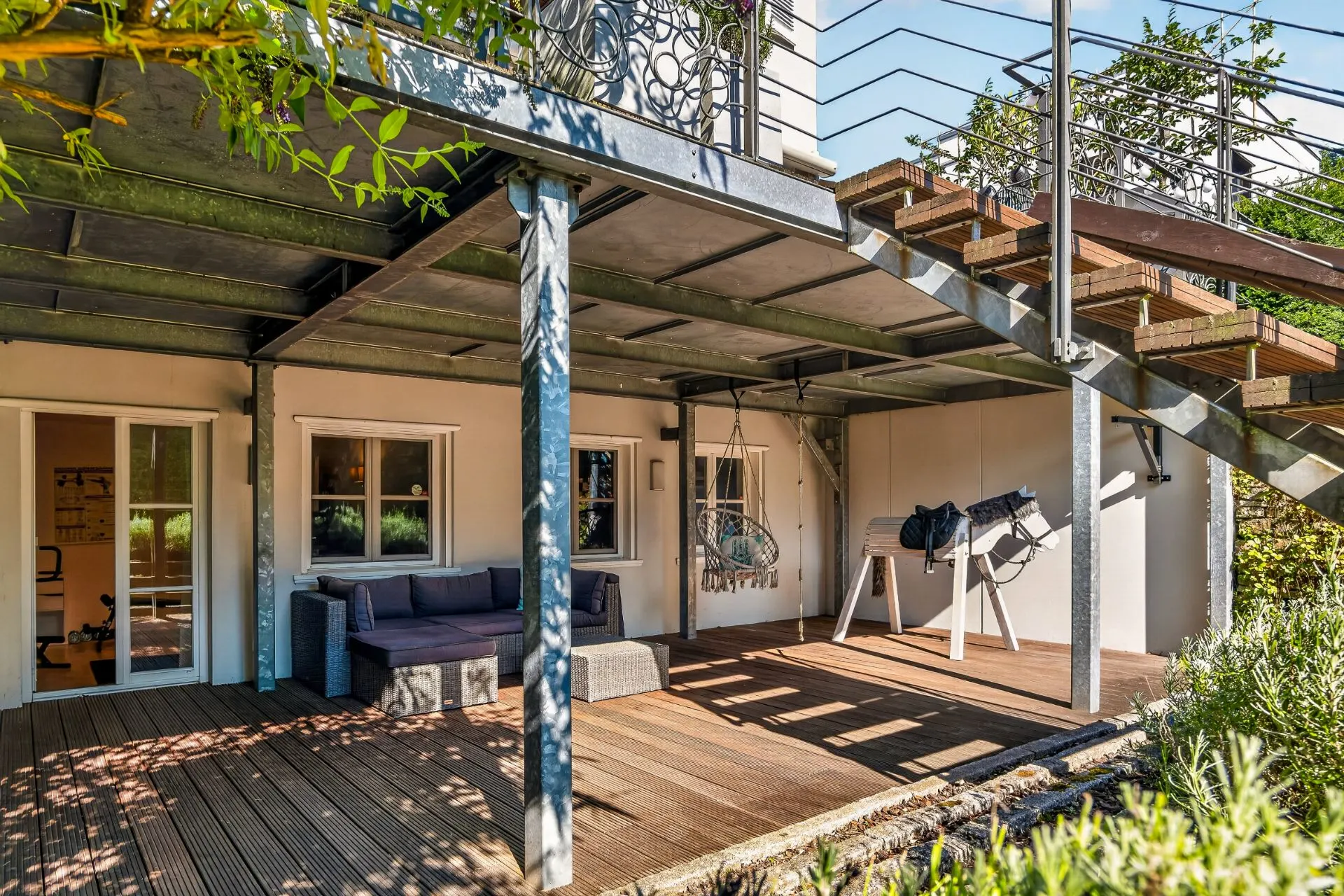 Überdachte Holzterrasse mit Sofa, Hängesessel und seitlicher Treppe an einem Wohnhaus.