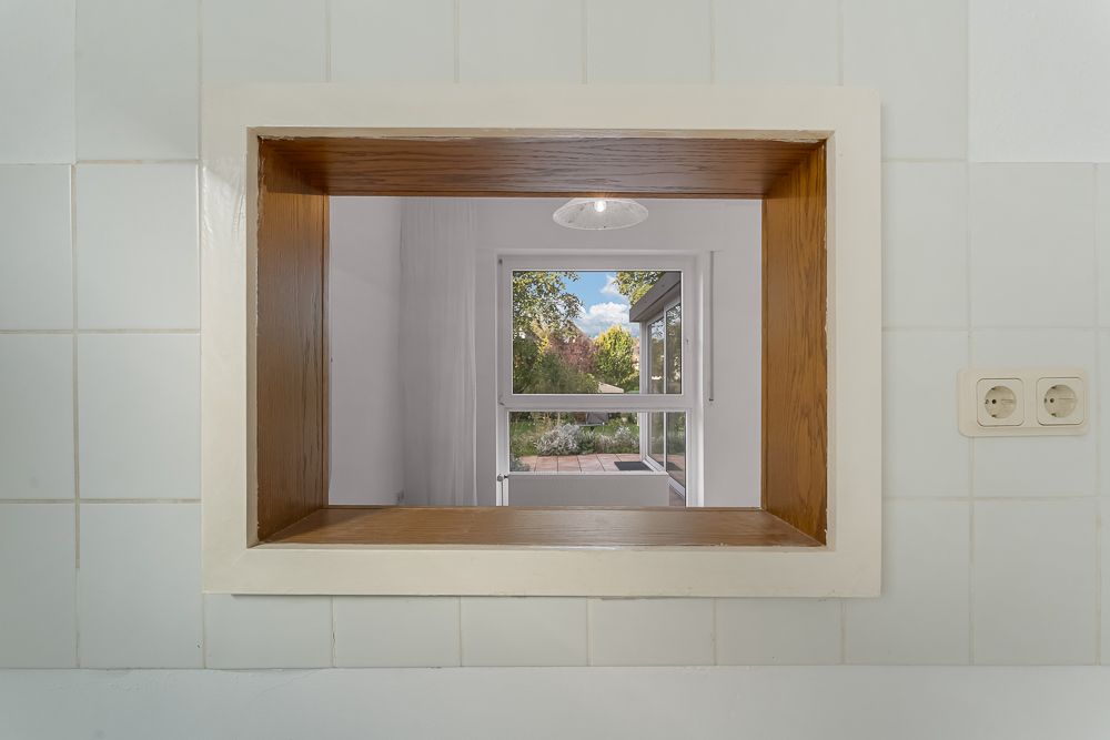 Durchreiche mit Holzrahmen in einer gefliesten Wand, dahinter Fenster mit Gartenblick.