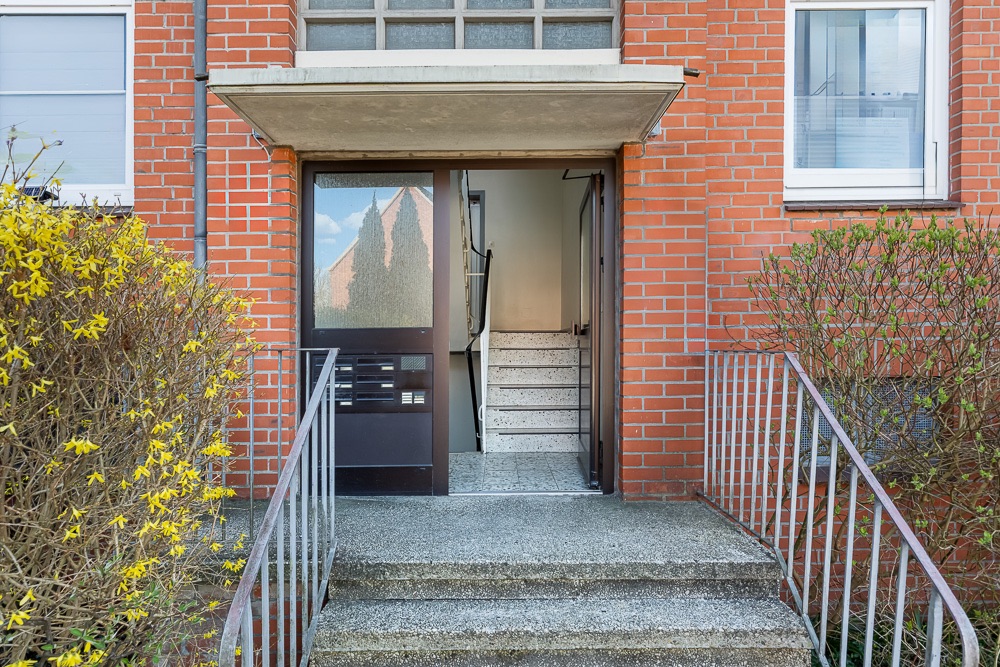 Eingang eines Mehrfamilienhauses mit Treppe und Glastür, umgeben von Büschen.
