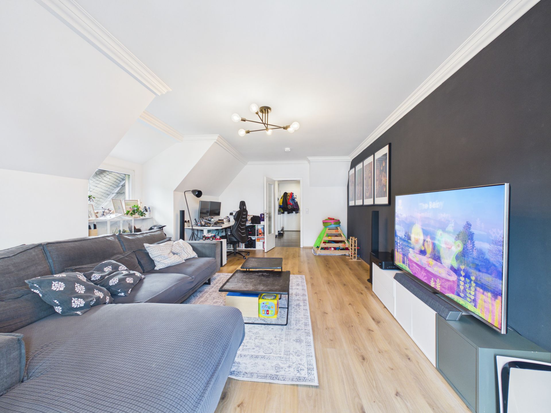 Modernes Wohnzimmer mit grauem Sofa, großem Fernseher, Schreibtisch und Holzfußboden.