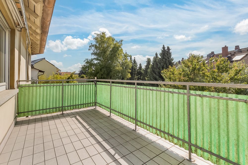 Großer Balkon mit grünem Sichtschutz und Fliesenboden, Ausblick auf Bäume und Nachbarhäuser.
