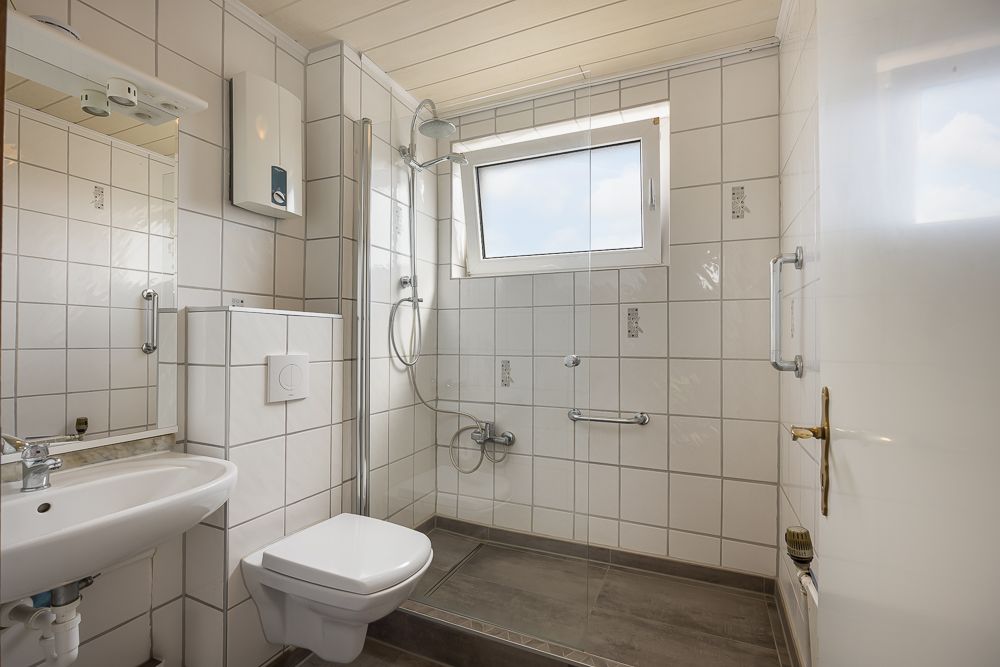 Modernes Badezimmer mit weiß gefliesten Wänden, ebenerdiger Dusche, Waschbecken und Fenster.