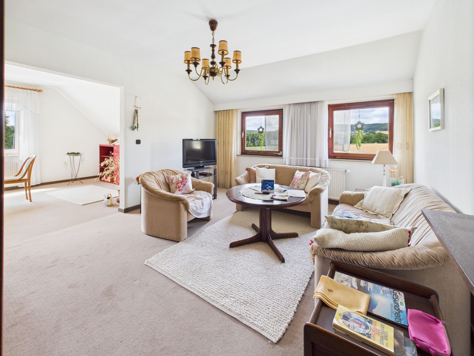 Helles Wohnzimmer mit Sofa, Sesseln, Couchtisch, Fernseher und zwei großen Fenstern.