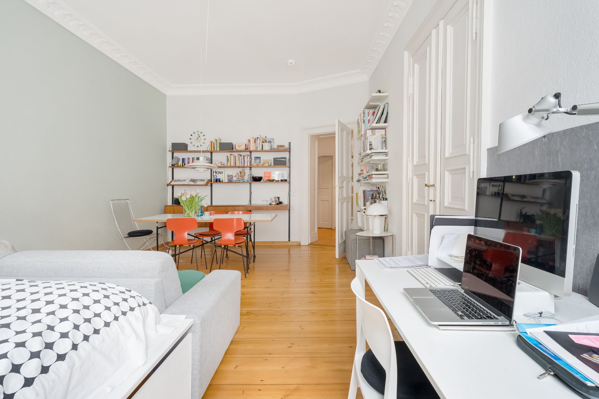 Helles Zimmer mit Schreibtisch, Sofa, Esstisch und Wandregalen mit Büchern.