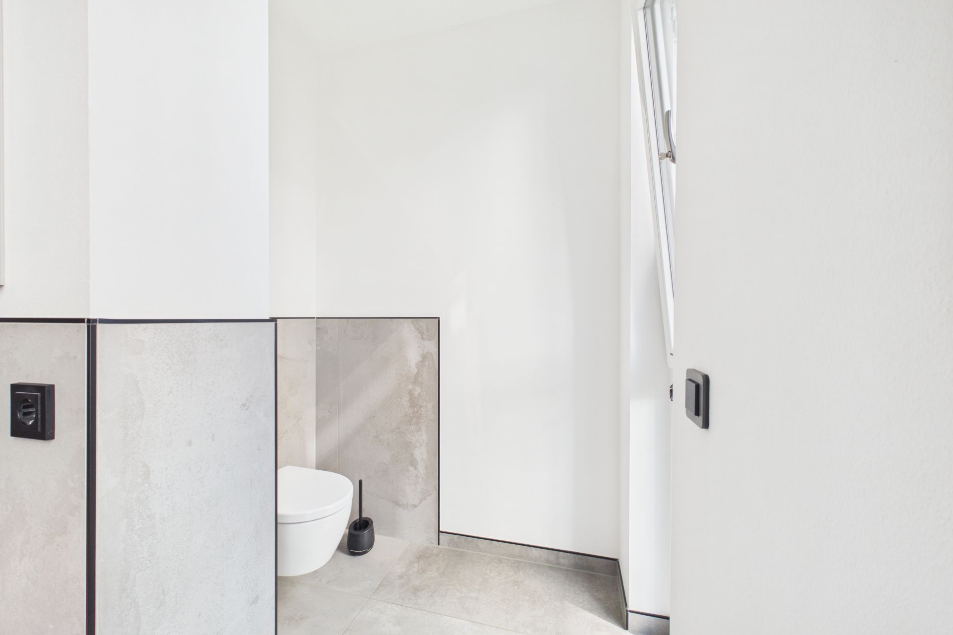 Helles Badezimmer mit Wandtoilette, Toilettenbürste und halb geöffnetem Fenster.