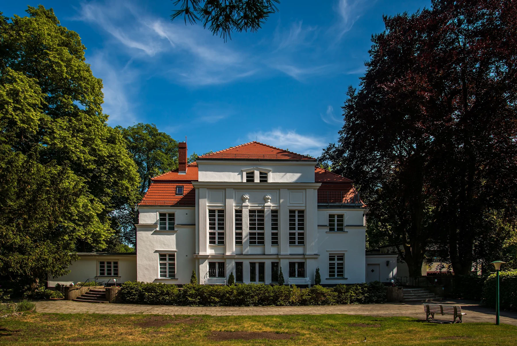 Großes weißes Wohnhaus mit rotem Dach, vielen Fenstern und Säulen, umgeben von Bäumen.