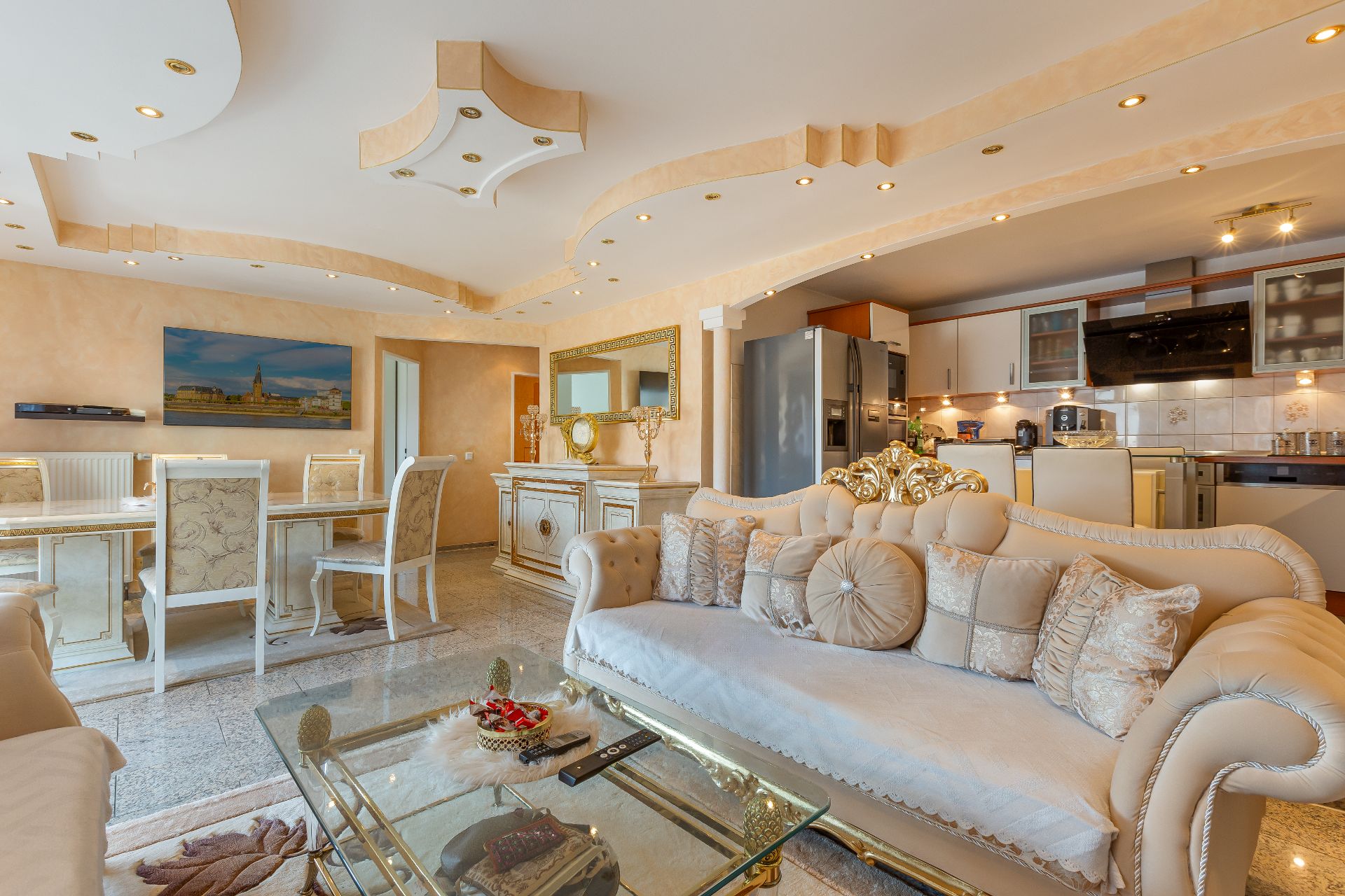 Helles Wohnzimmer mit eleganter Sofagarnitur, Esstisch, Glastisch und moderner Küche im Hintergrund.