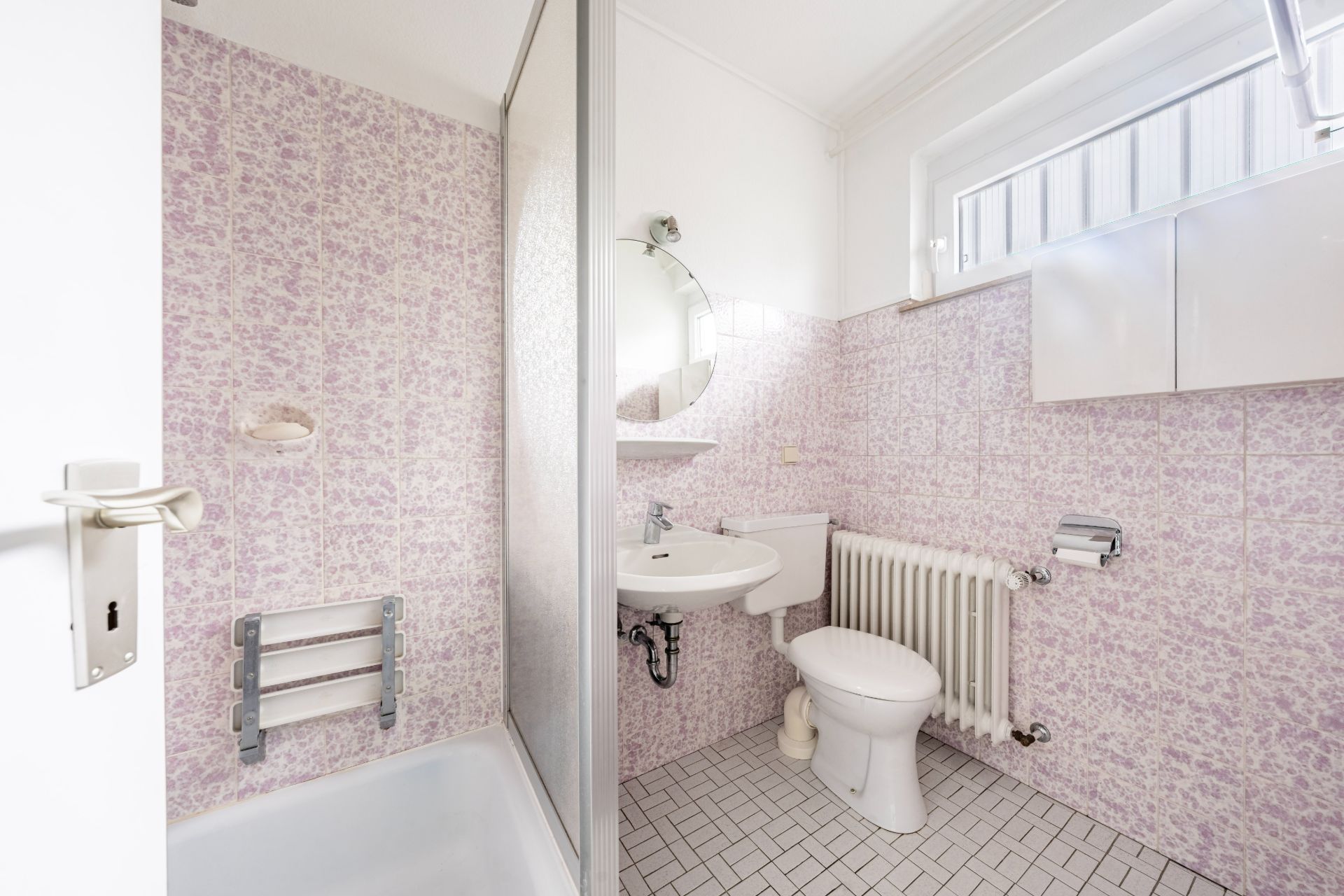 Kleines Badezimmer mit Dusche, Waschbecken, Toilette, Heizung und Fenster.