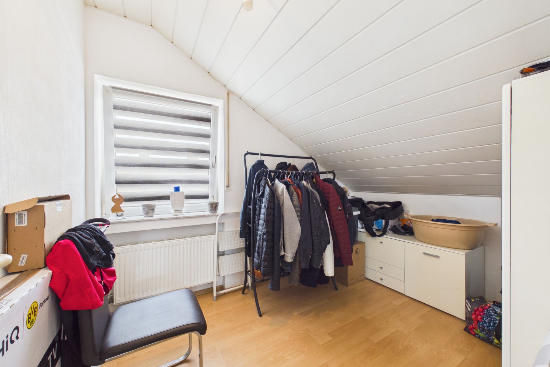 Kleines Zimmer mit Schräge, Kleiderständer, Stuhl, Kommode und Fenster mit Rollos.