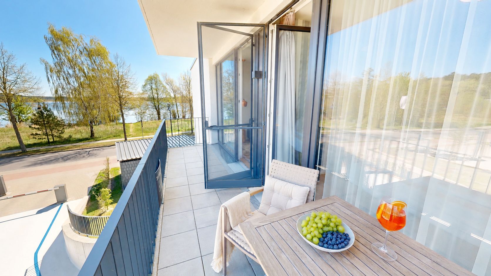 Heller Balkon mit Tisch, Stuhl, einem Obstteller und einem Getränk, Blick auf Bäume und See.
