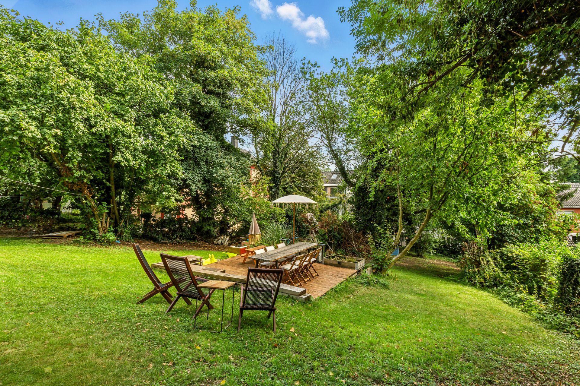Garten mit hölzener Terrasse, langen Tischen, Stühlen und Sonnenschirm, umgeben von Bäumen.