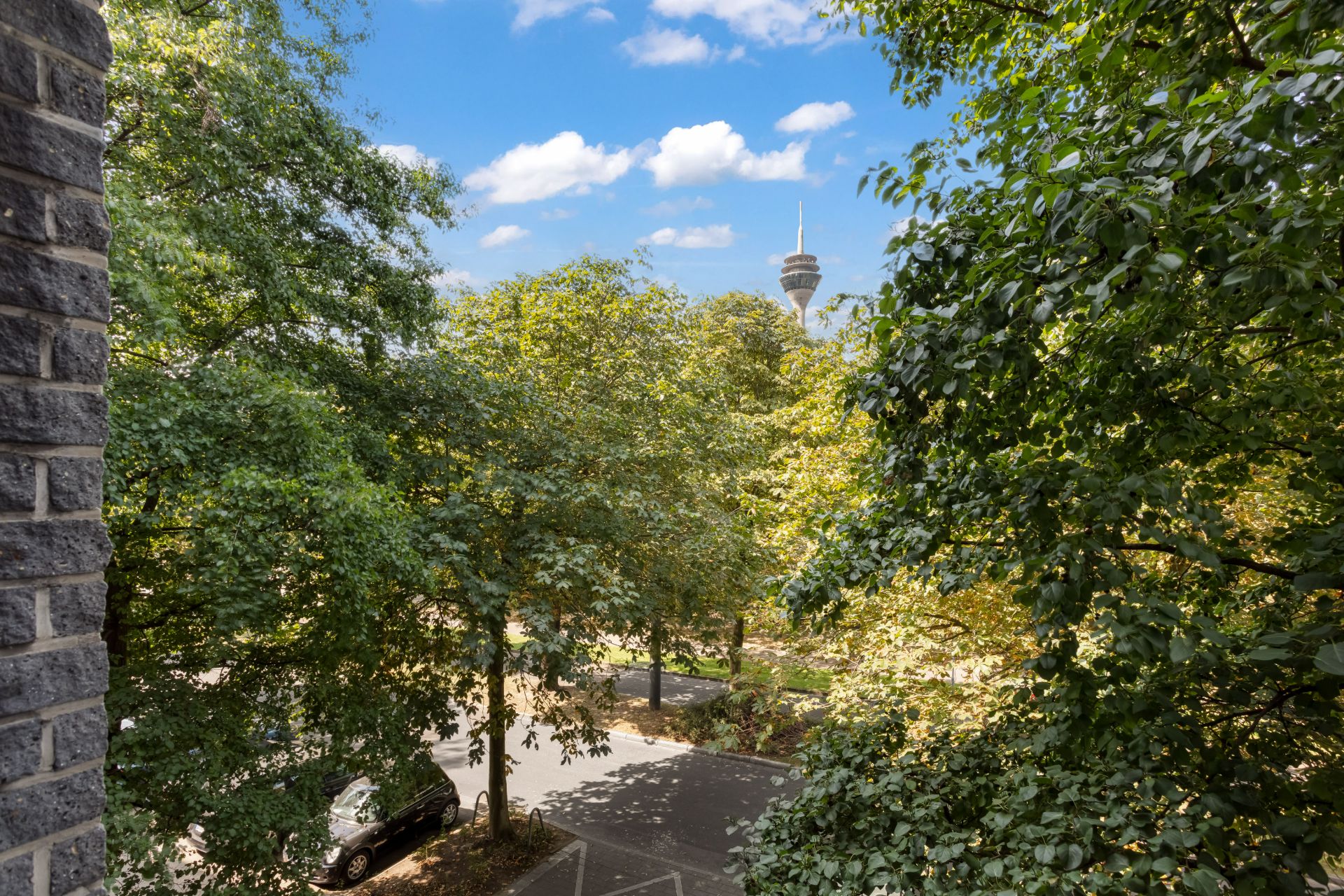 Blick aus dem Fenster auf eine von Bäumen gesäumte Straße mit Autos und Fernsehturm im Hintergrund.