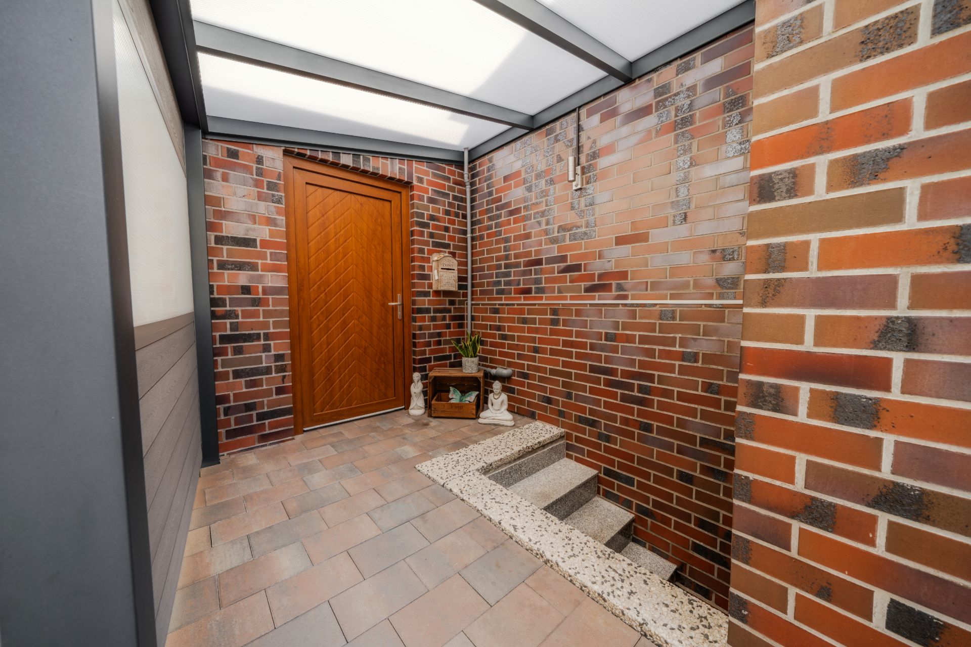 Überdachter Eingangsbereich mit brauner Tür, Backsteinwänden und kleiner Treppe.