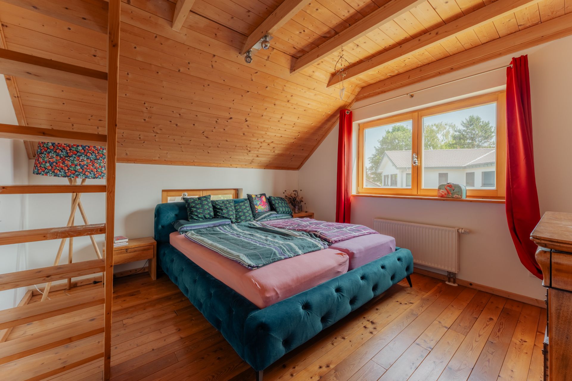 Schlafzimmer mit Holzboden, Doppelbett, großem Fenster und rotem Vorhang unter schräger Holzdecke.