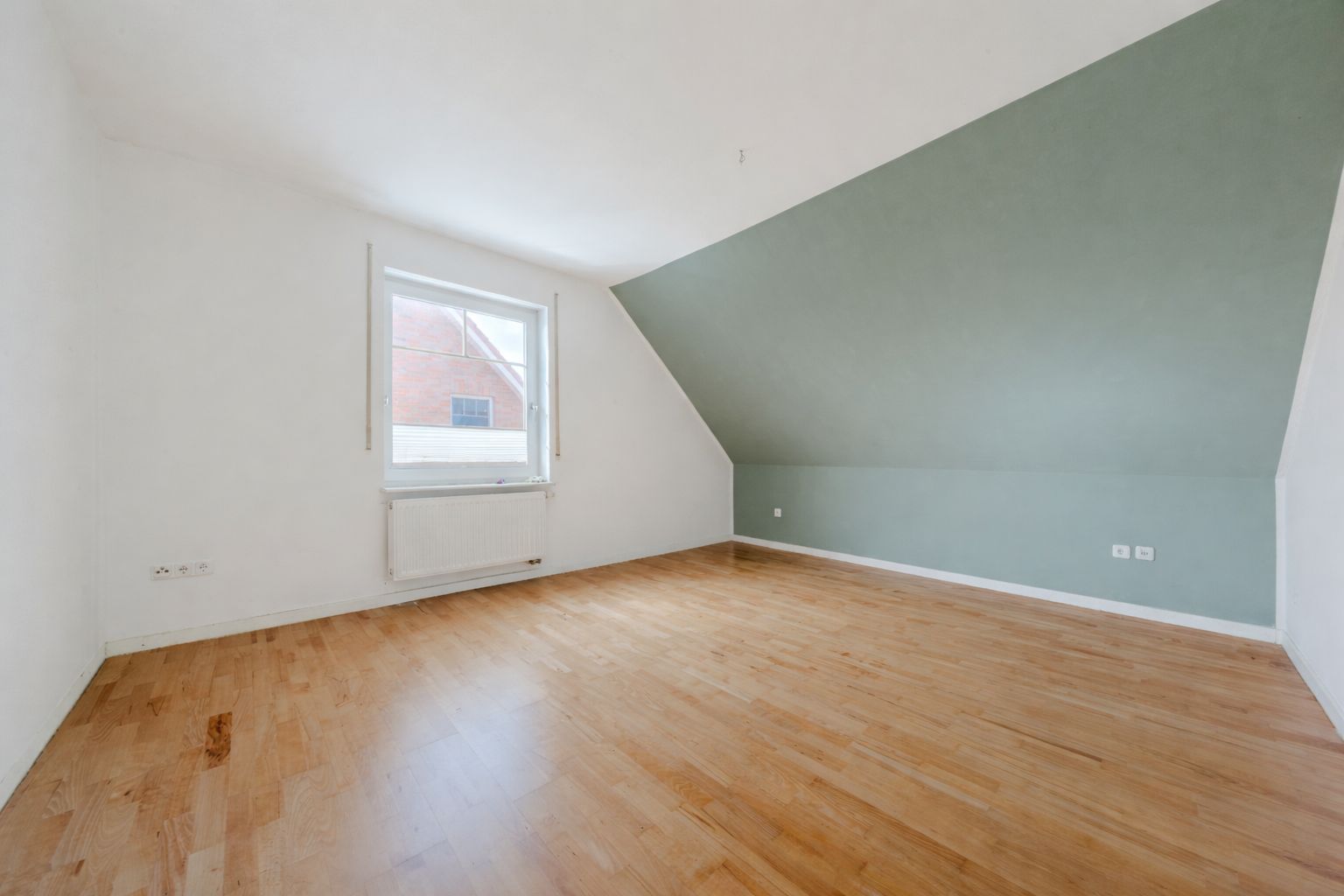 Leeres Zimmer mit Holzboden, einem Fenster und einer schrägen, grün gestrichenen Wand.
