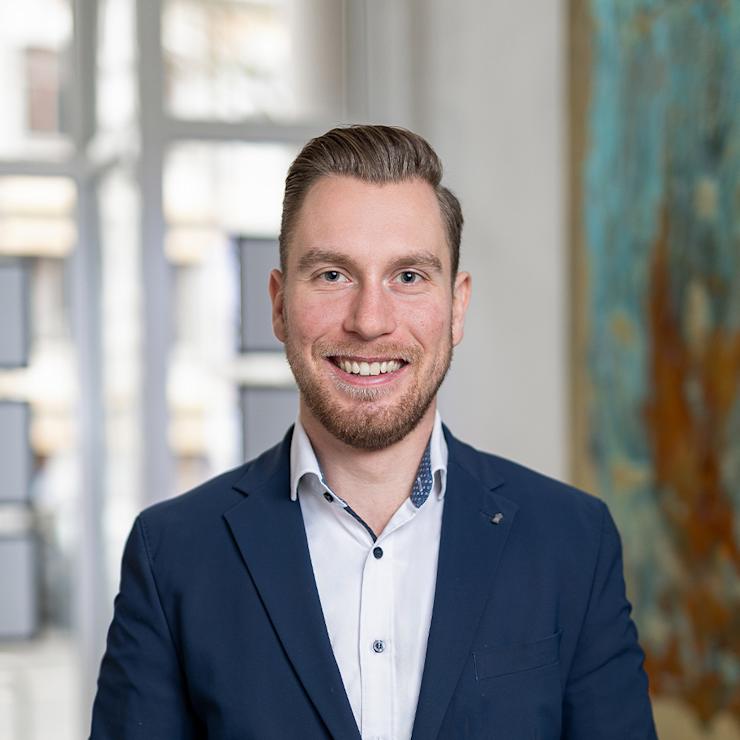 Ein Mann in einem blauen Anzug steht lächelnd in einem hellen Raum vor einem Fenster.