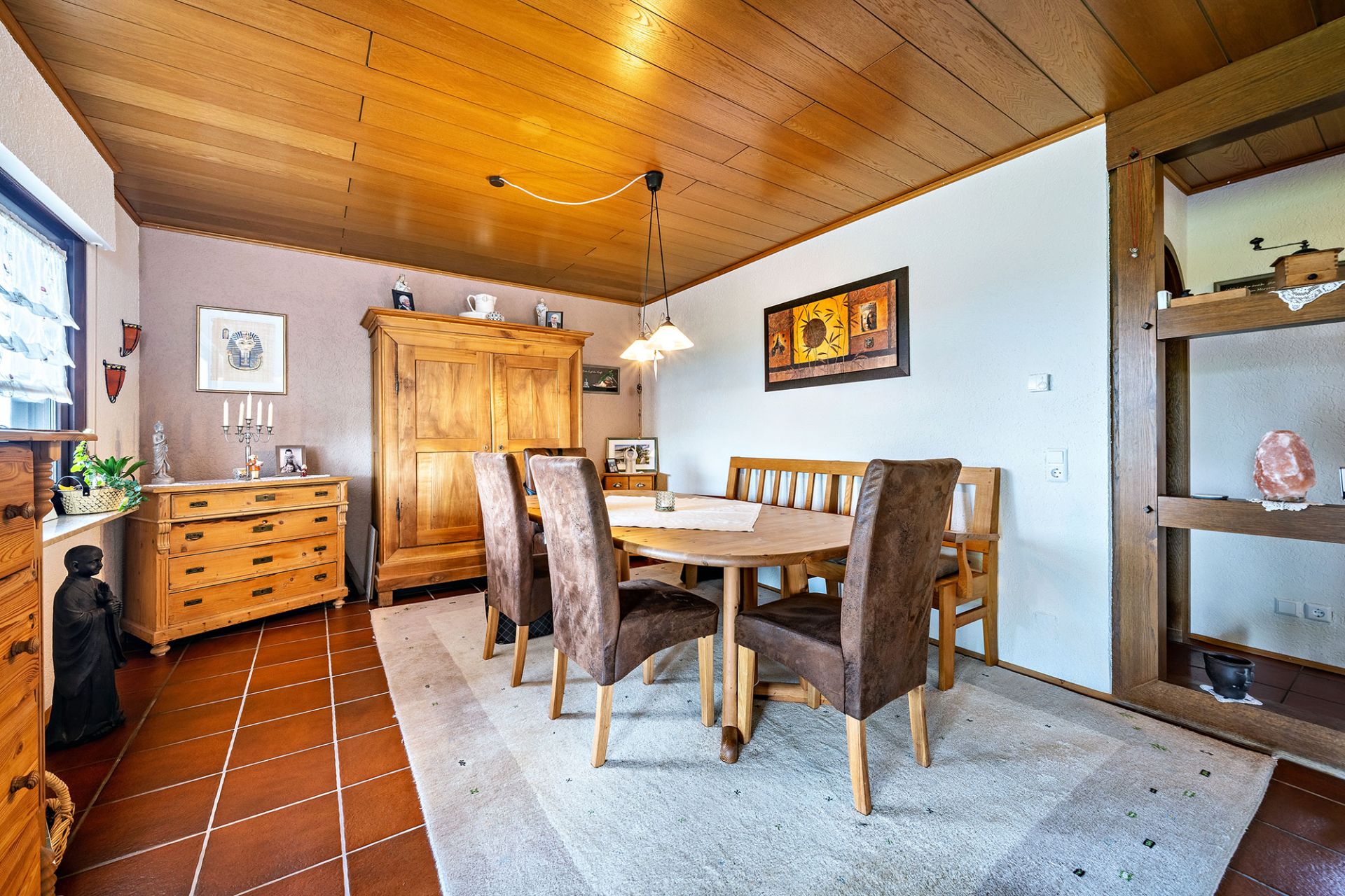 Esszimmer mit Holzmöbeln, rundem Tisch, braunen Stühlen und Schrank auf Fliesenboden.