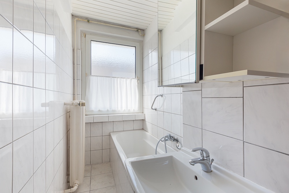 Helles, schmales Badezimmer mit Badewanne, Waschbecken und Fenster mit Vorhang.