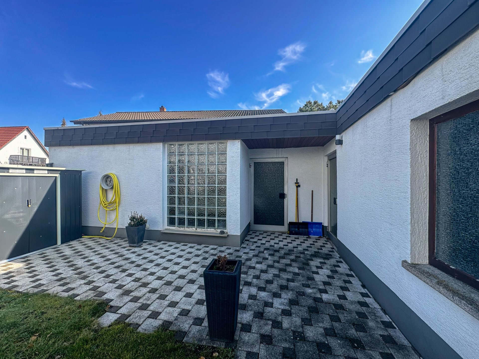 Gepflasterte Terrasse mit Pflanzkübel, Gartenschlauch und Glasbausteinfenster an einem weißen Haus.