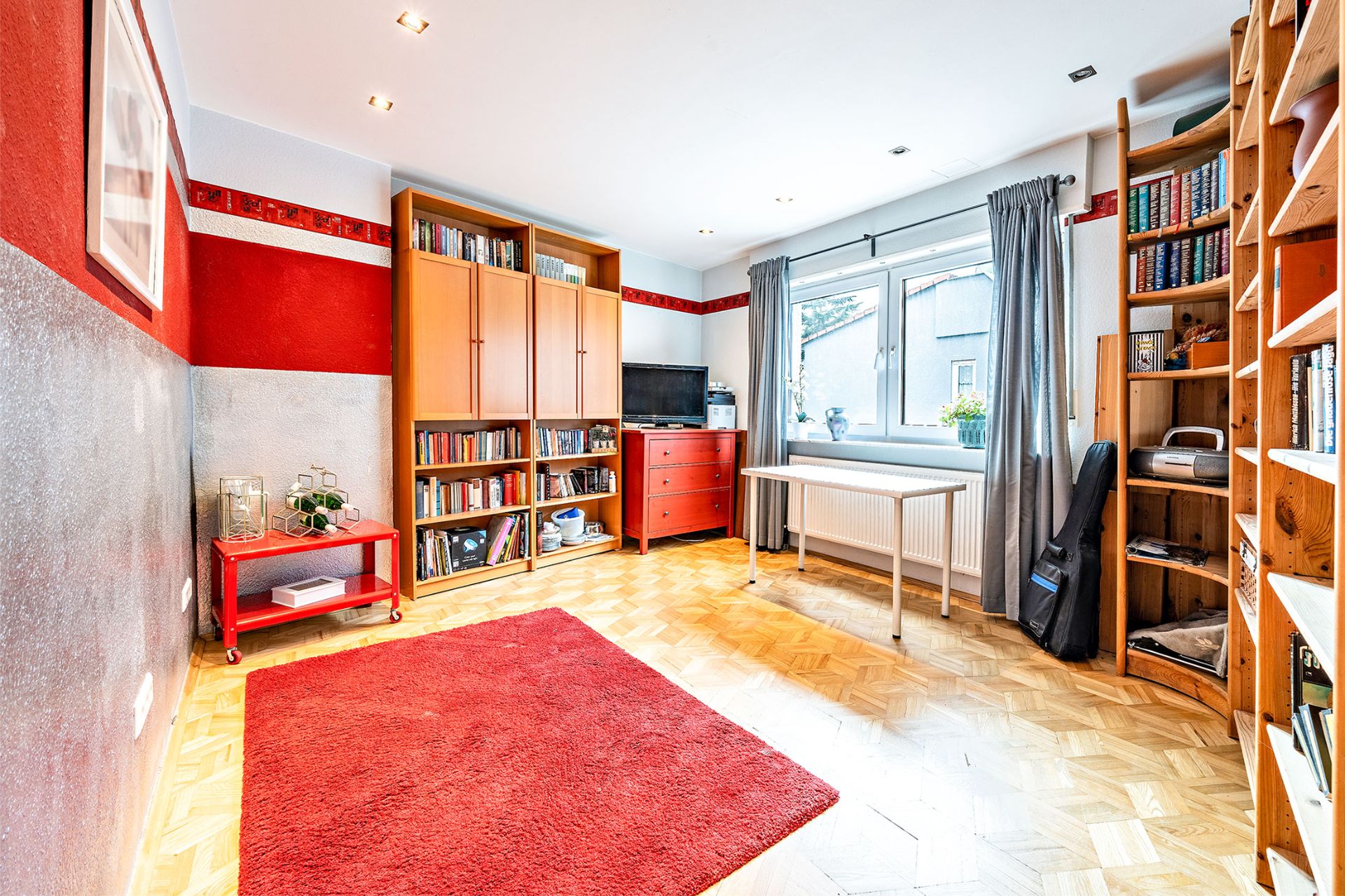 Helles Zimmer mit Bücherregalen, rotem Teppich und großem Fenster mit Vorhängen.