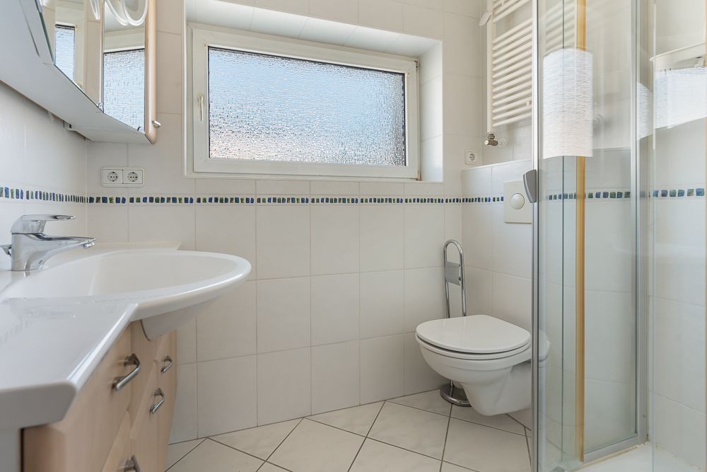 Helles Badezimmer mit Waschbecken, Toilette, Fenster und Duschkabine.