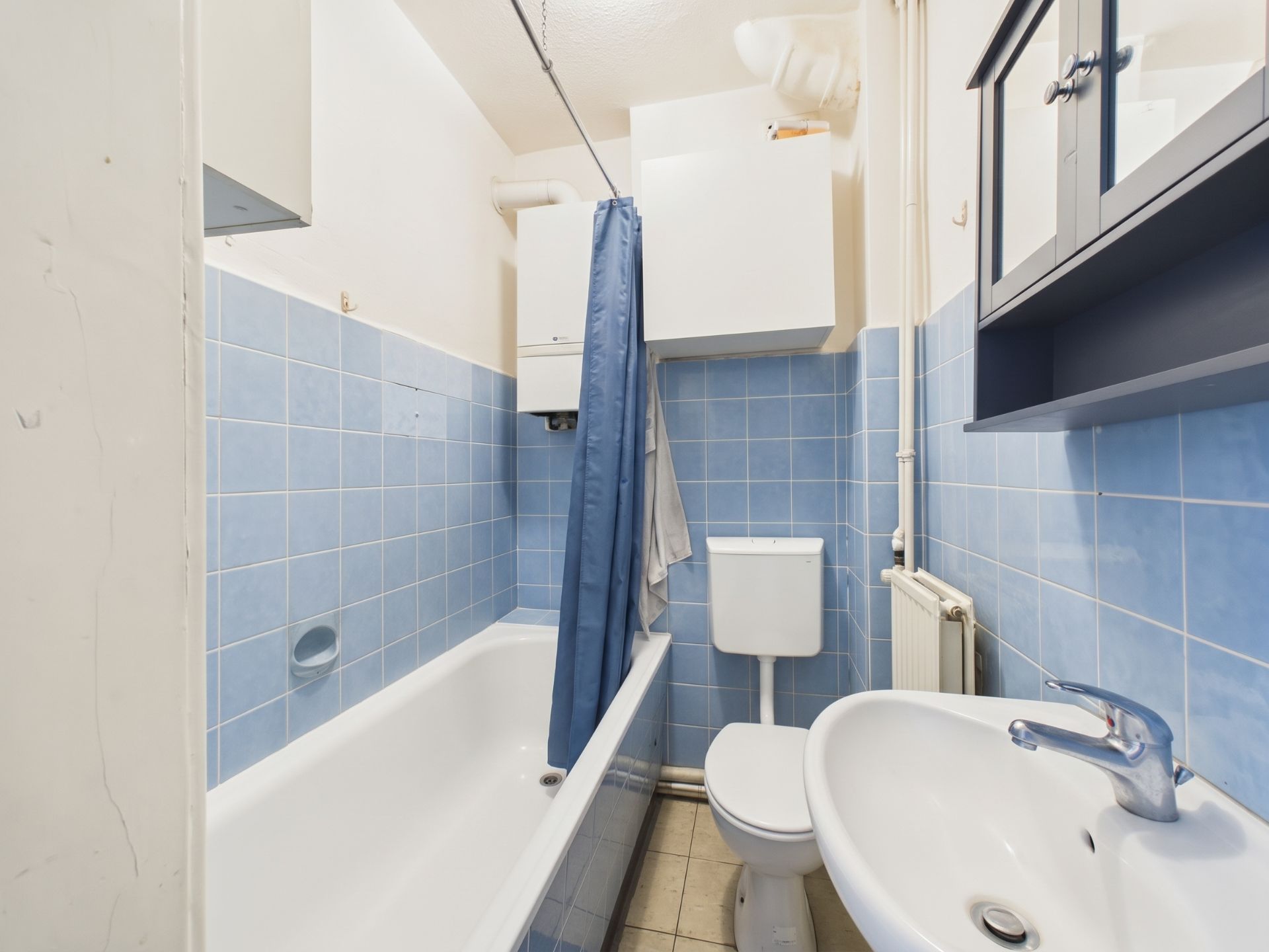Kleines Badezimmer mit Badewanne, blau gefliesten Wänden, Waschbecken, Toilette und Spiegelschrank.