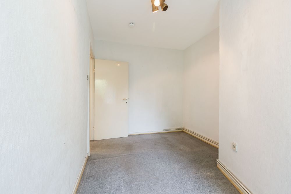 Schmaler, leerer Raum mit Teppichboden, weißen Wänden und geöffneter Tür.
