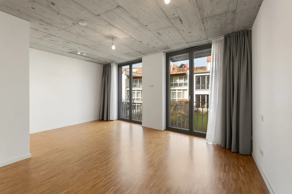 Leerer Raum mit Holzboden, großen Fenstern und grauen Vorhängen, Blick auf einen Innenhof.