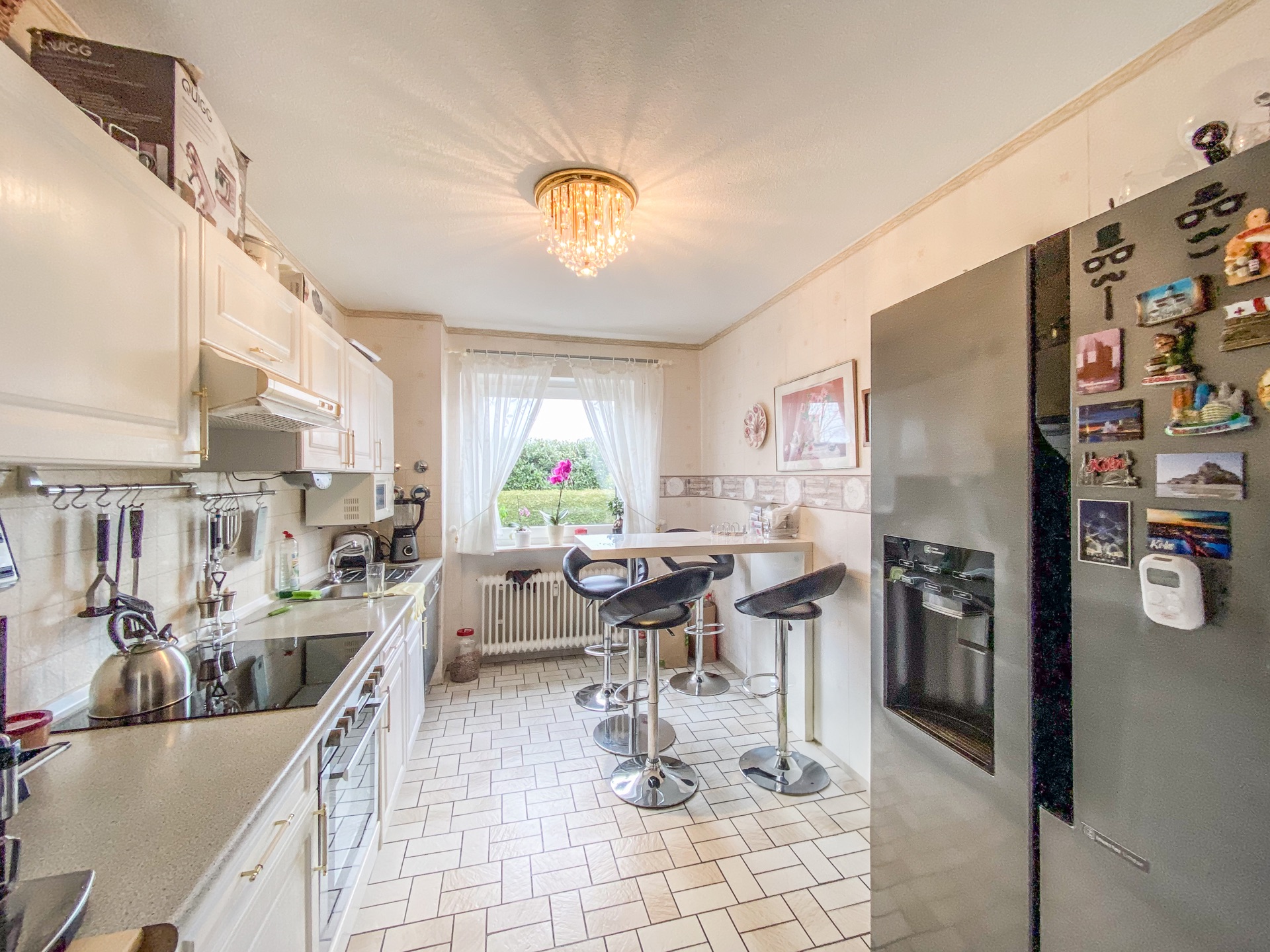 Helle Küche mit weißen Schränken, Kühlschrank, Bartisch, drei Hockern und Fenster mit Vorhängen.